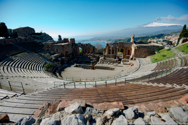 Curving theatre seating surrounds the stage ruins, with wooden seating surfaces in front and Mount Etna beyond.