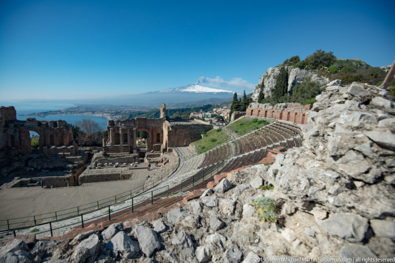 Ancient theatre seating and stage ruins below rocky masonry, with snow-covered Mount Etna on the distant horizon.