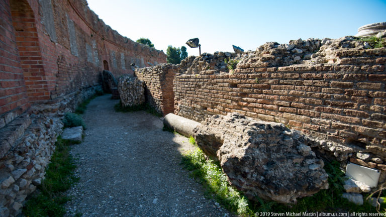 Narrow gravel passage between weathered brick and stone walls, with a large fallen masonry fragment beside the path.