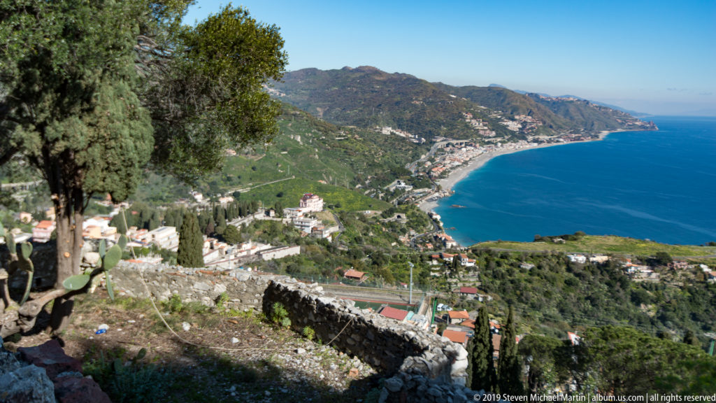 Stone remnants and cactus on a high slope overlooking coastal towns, mountains, and the blue Ionian Sea.