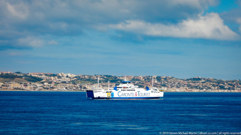 White Caronte & Tourist ferry crossing blue water in front of a densely developed hilly shoreline.