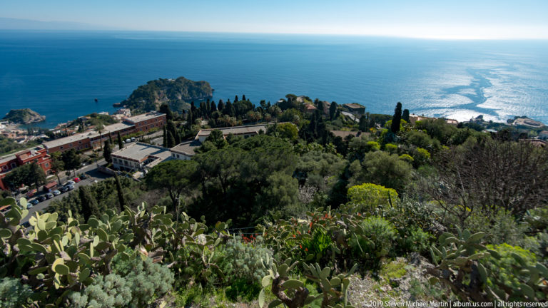 Green hillside with cypress and prickly pear descending toward a small promontory surrounded by the blue Ionian Sea.