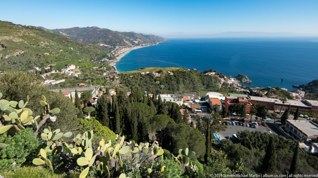 Prickly pear cactus and trees above a curving blue coastline backed by steep green mountains and coastal towns.