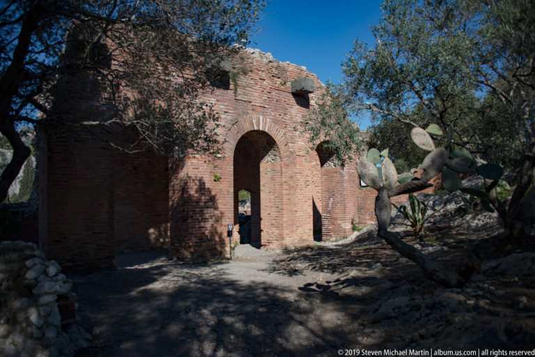 Weathered brick ruin with a tall central arch, smaller openings, olive trees, and prickly pear under a clear blue sky.