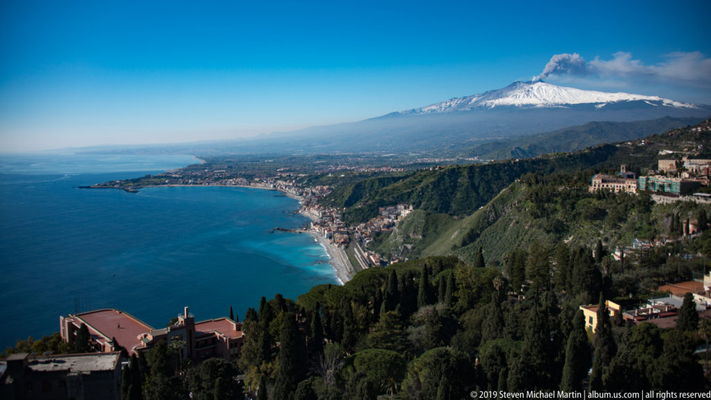 Snow-covered Mount Etna beyond a steep green coastline and blue Ionian Sea, viewed from high above Taormina.