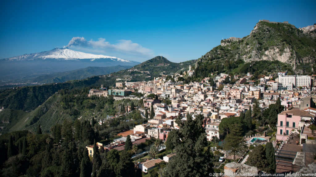 Dense Taormina hillside buildings and rocky peaks in the foreground with snow-covered Mount Etna across the distant horizon.