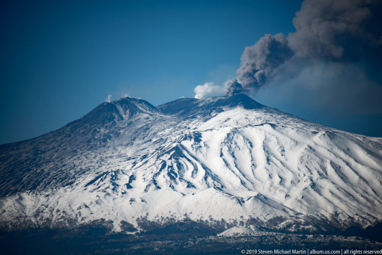 Snow-covered Mount Etna with dark volcanic ridges and a dense gray plume rising from the summit into blue sky.