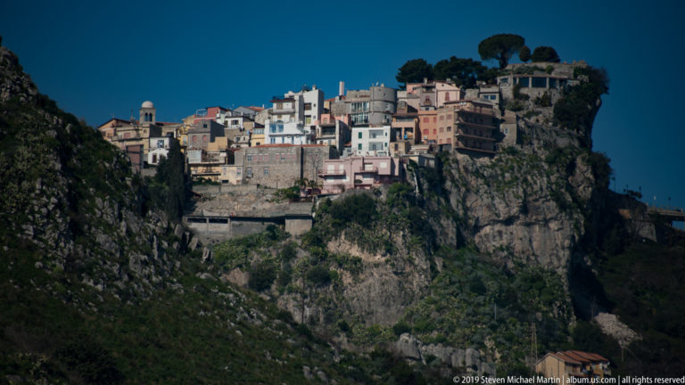 Pastel and pale buildings perched atop a sheer rocky cliff above a deep green ravine under clear blue sky.