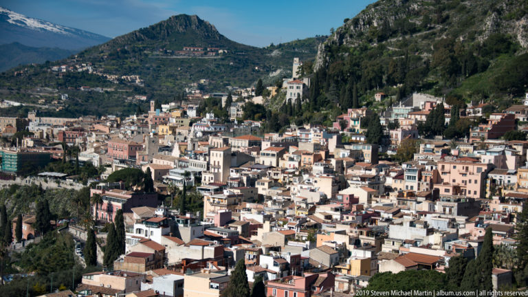 Dense Taormina townscape of pale buildings, church towers, cypress trees, and steep rocky green slopes.