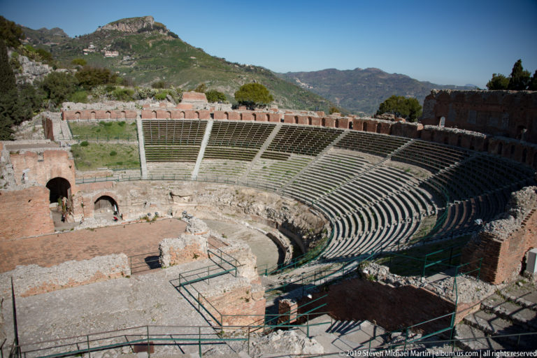 Wide semicircular ancient theatre seating with pale radial stairs, brick perimeter walls, and green hills beyond.