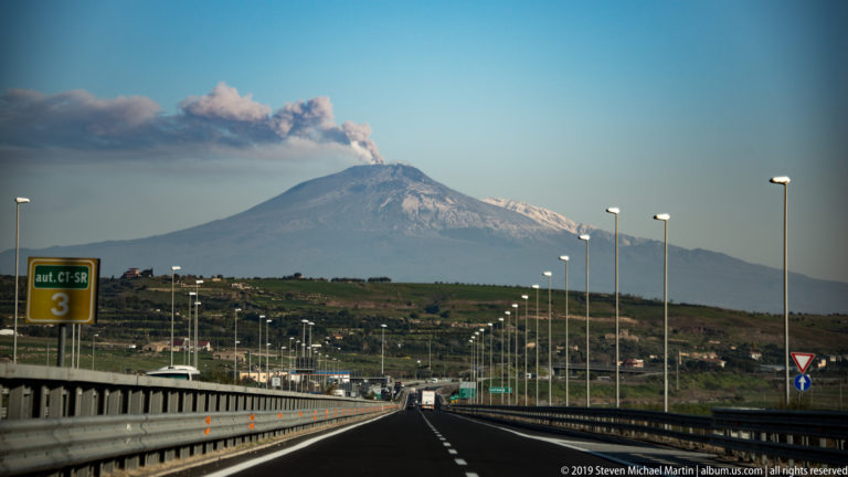 Straight motorway with rows of streetlights leading toward Mount Etna, where a plume trails from the snow-streaked summit.