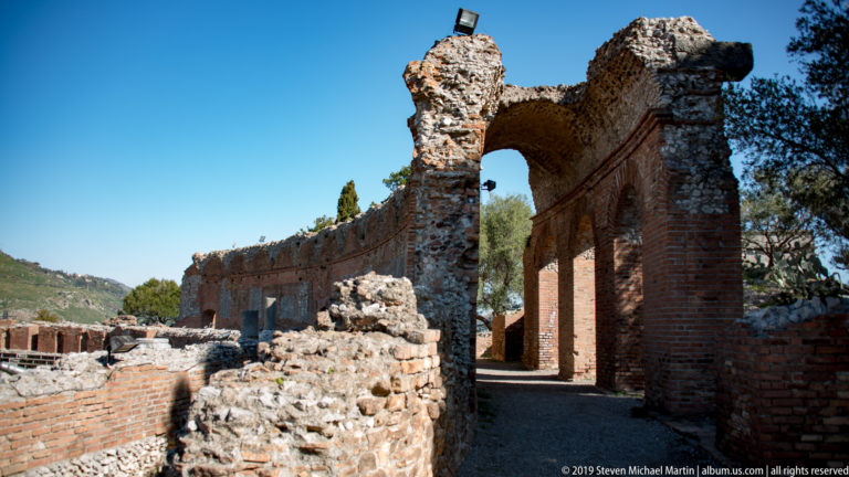Long weathered brick arcade with repeated arches, ruined wall tops, rubble masonry, and a narrow path through the ancient theatre.