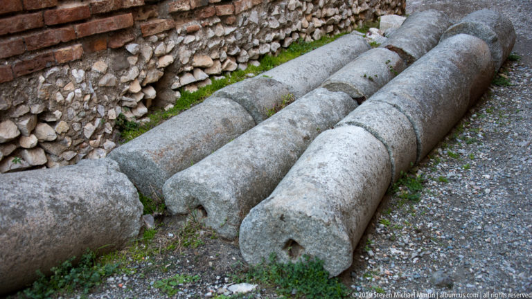 Three large weathered stone column shafts lying parallel on the ground beside an ancient rubble wall.