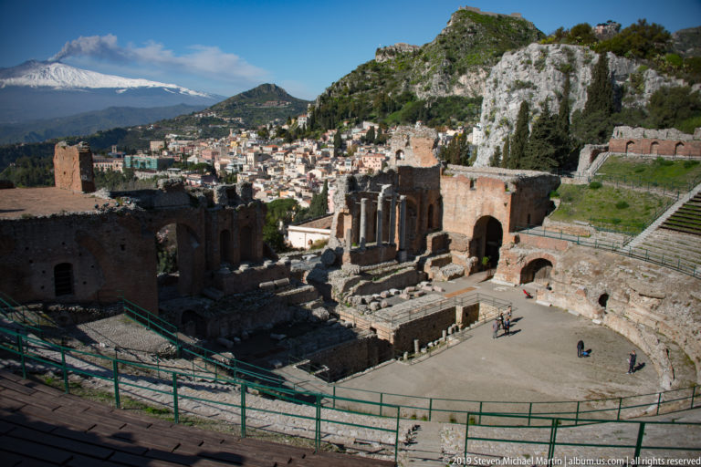 Wide view of ancient theatre seating, performance area, stage ruins, Taormina buildings, and rocky hills under blue sky.