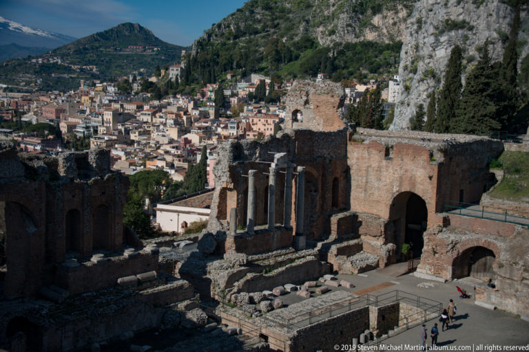 Ancient theatre stage ruins and standing columns with dense Taormina buildings and a steep limestone ridge behind.