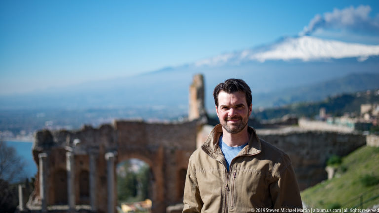 Smiling man in tan jacket with blurred brick theatre ruins and a broad view of snowy Mount Etna behind.