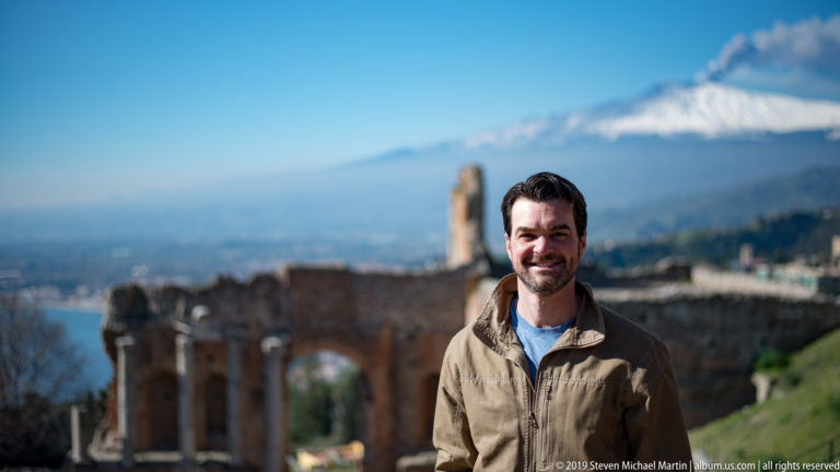 Smiling man in tan jacket before blurred ancient theatre ruins and snow-covered Mount Etna.