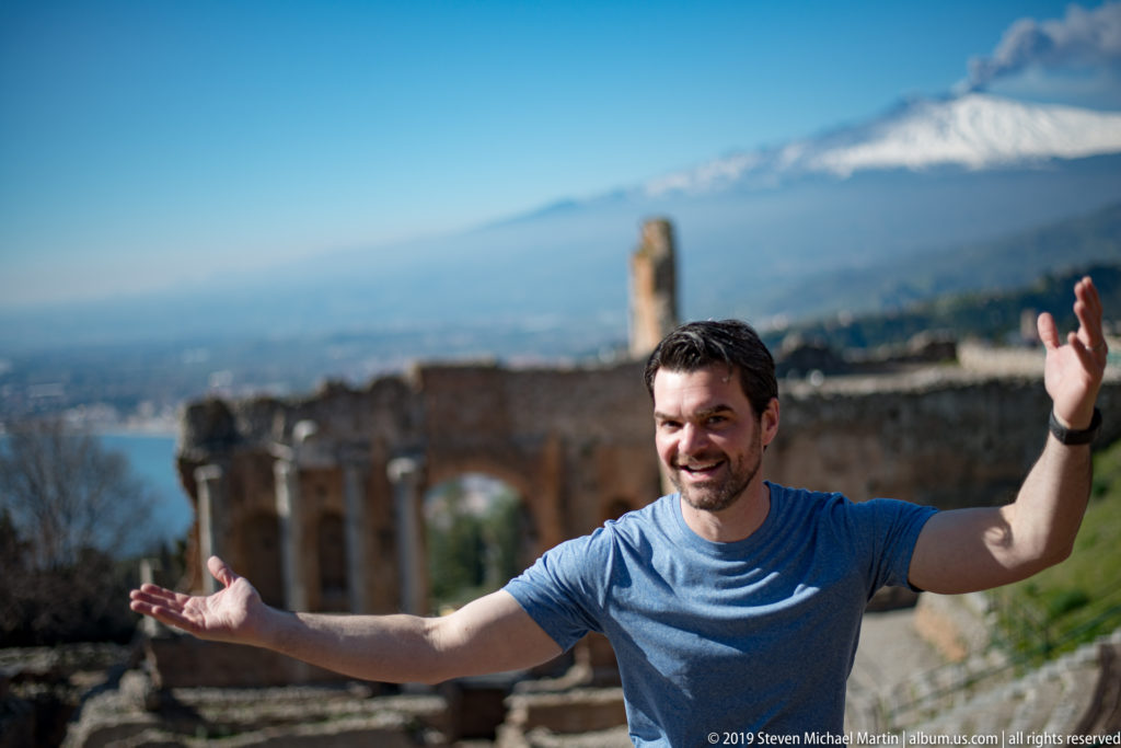 Smiling man in blue shirt with arms spread wide before ancient theatre ruins and snow-covered Mount Etna.