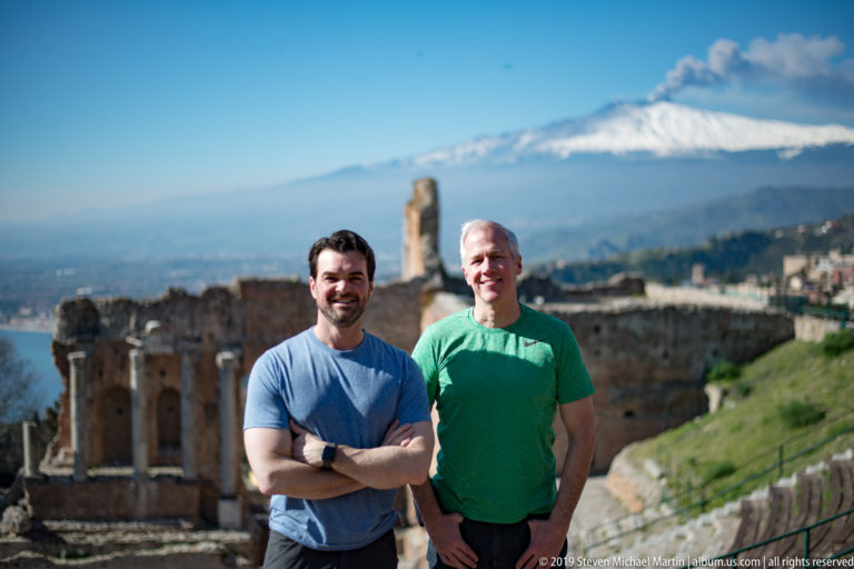 Two smiling men in blue and green shirts posed together with ancient theatre ruins and snowy Mount Etna behind.