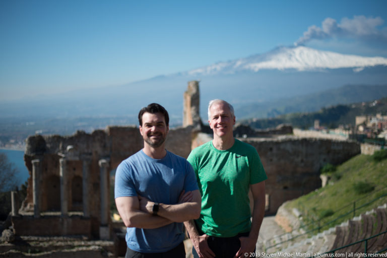 Two smiling men in blue and green shirts standing before ancient theatre ruins and snow-covered Mount Etna.