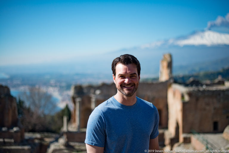 Close portrait of a smiling man in blue with blurred ancient theatre ruins and snow-covered Mount Etna behind.