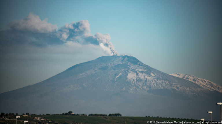 Distant Mount Etna above dark fields, with patchy snow and a compact plume rising from the summit.