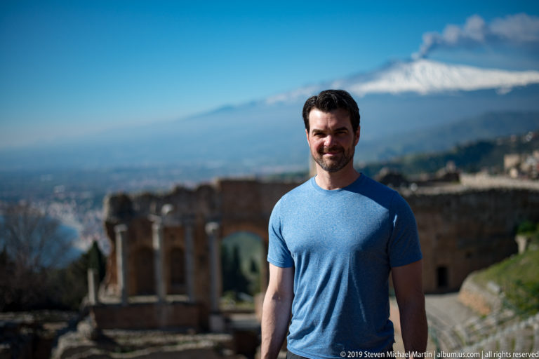 Smiling man in blue shirt standing before ancient theatre ruins with snow-covered Mount Etna in the background.