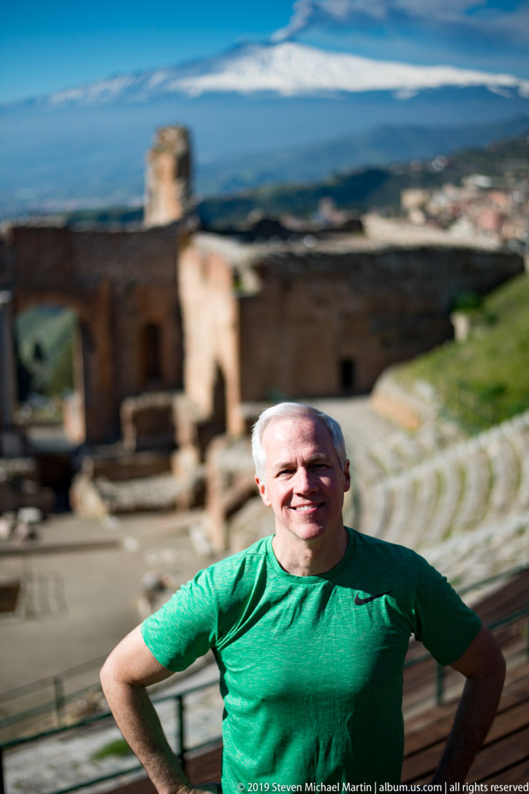 Smiling man in green shirt with curving ancient theatre seating, stage ruins, and snow-covered Mount Etna behind him.
