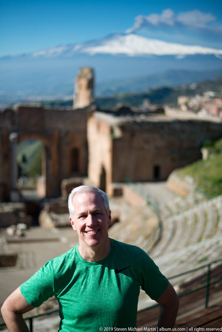 Smiling man in a green shirt before blurred ancient theatre ruins and snow-covered Mount Etna.