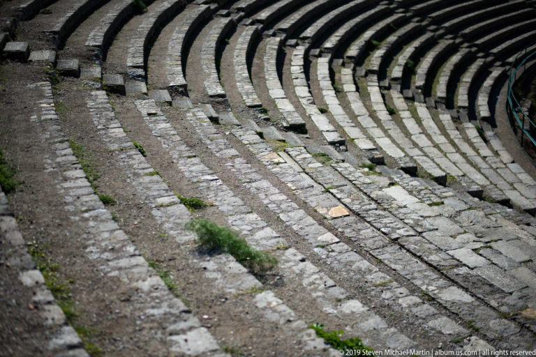 Diagonal view across curved stone seating tiers and steps, with sparse grass growing between weathered blocks.