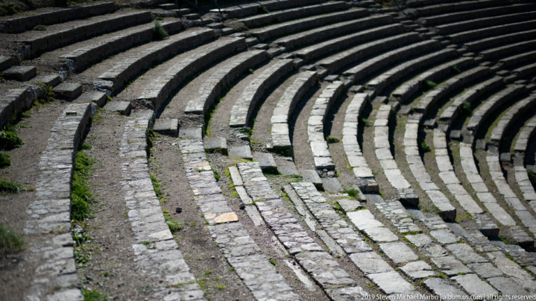 Curving rows of weathered stone theatre seats and narrow steps with small plants growing between masonry joints.