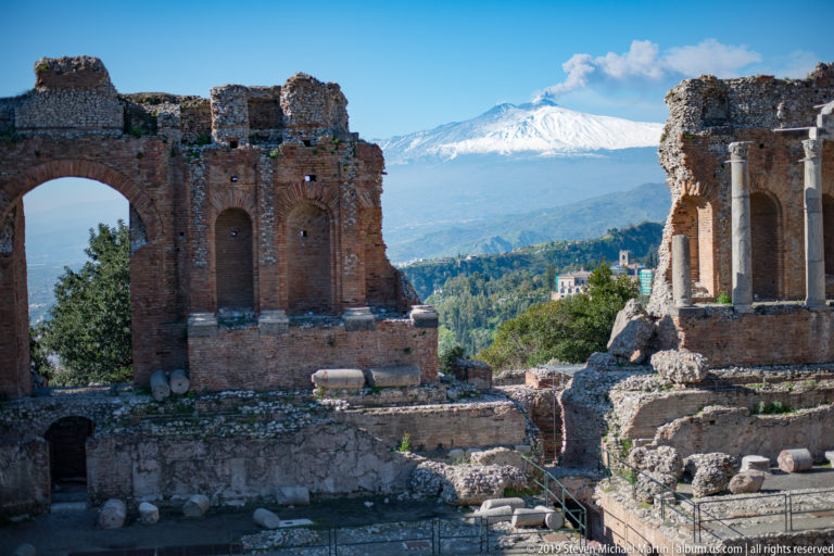 Brick stage ruins, standing columns, and excavated foundations with snow-covered Mount Etna centered in the distance.