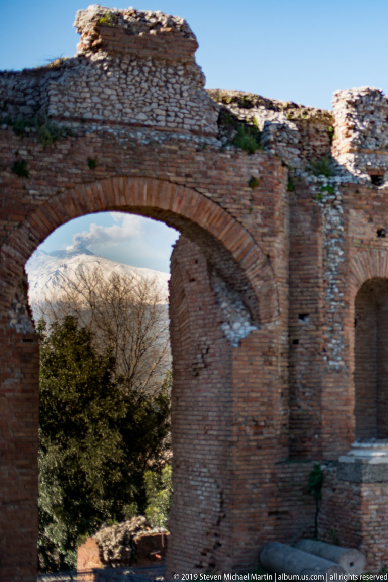 Weathered brick arch and rubble masonry with trees and snow-covered Mount Etna visible through and above the opening.
