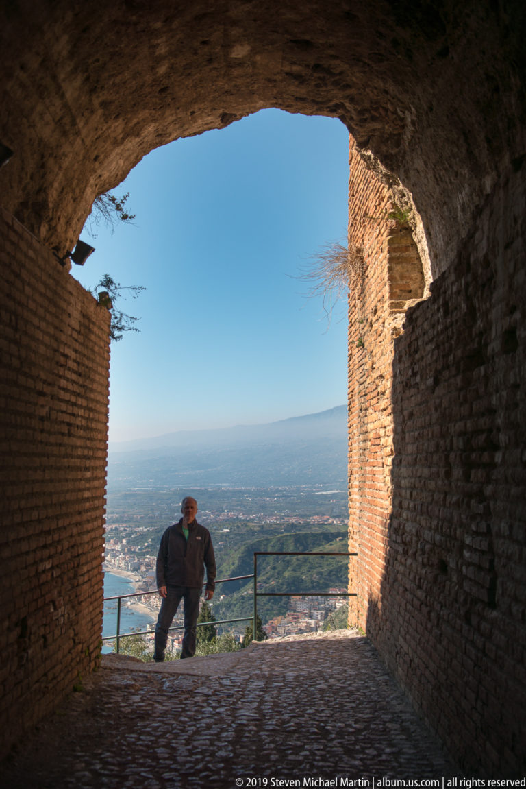 Visitor standing in a shadowed brick archway overlooking a sunlit coastal landscape and distant blue sea.