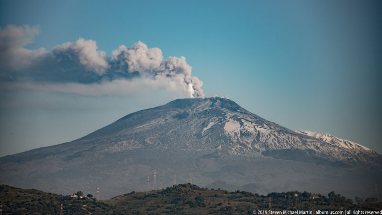 Mount Etna’s dark broad cone with patchy summit snow and a long gray plume drifting sideways across the sky.