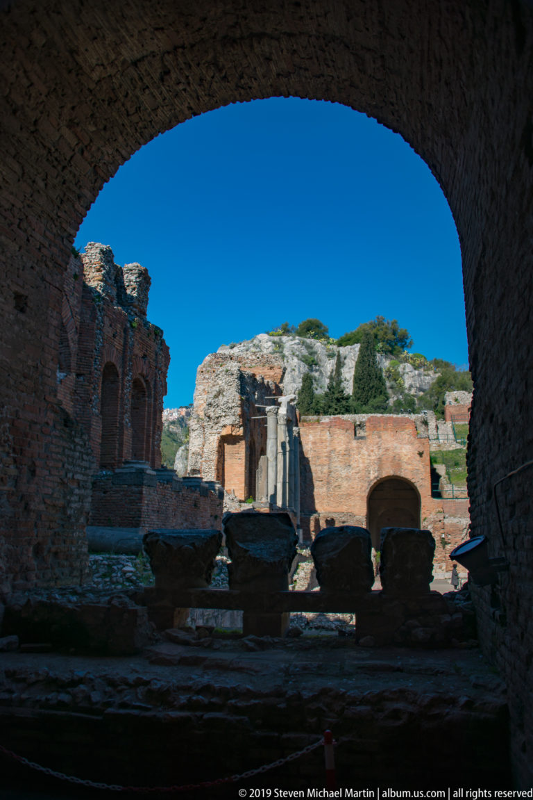 Ancient brick ruins, columns, and stone fragments in sunlight, framed by a broad dark vault with rocky hillside beyond.