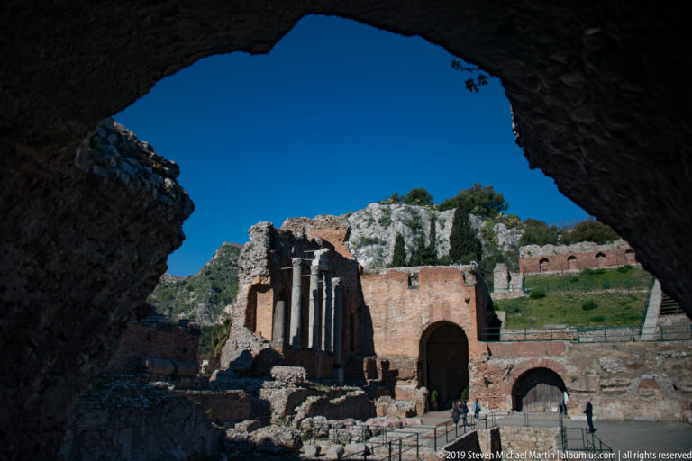 Sunlit ancient theatre ruins and standing columns seen through a large dark masonry opening, with rocky hillside beyond.