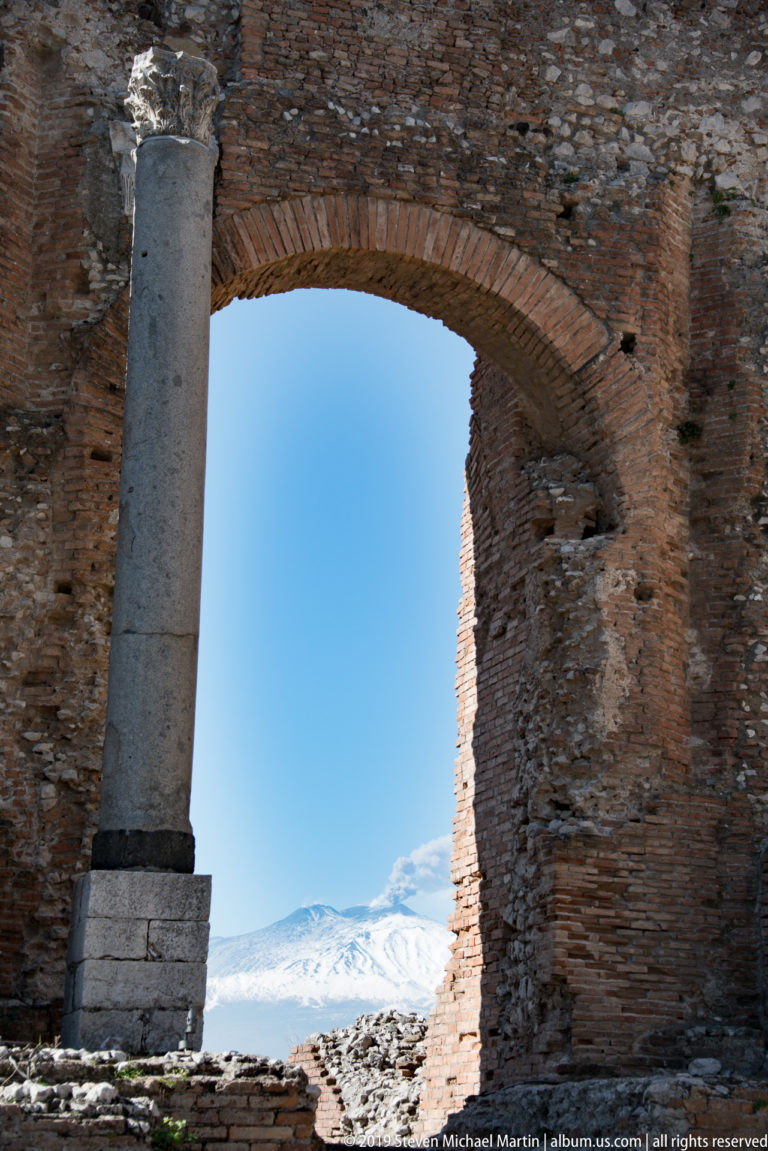 Large ancient brick arch and pale stone column framing the snow-covered slopes of Mount Etna beneath blue sky.