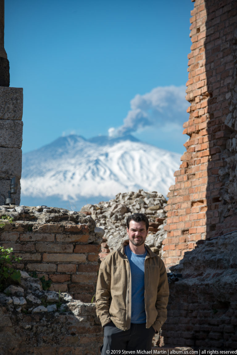 Man in a tan jacket standing among ancient brick ruins with snow-covered Mount Etna across the background.