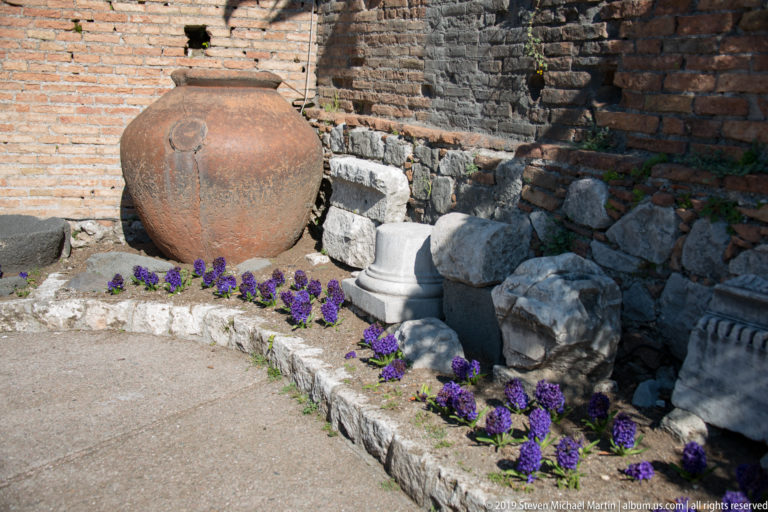 Large reddish ceramic storage vessel beside carved stone bases, rubble, brick walls, and purple flowers at the theatre.