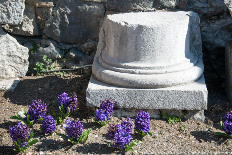 Weathered pale stone column base on a square plinth surrounded by small purple flowers and rough archaeological masonry.