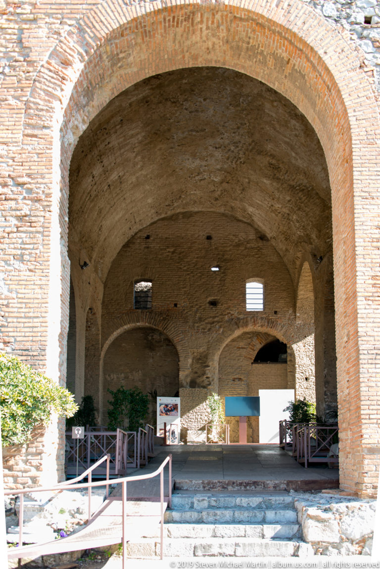 Large brick arch leading into a barrel-vaulted ancient interior with smaller openings, railings, steps, and visitor panels.