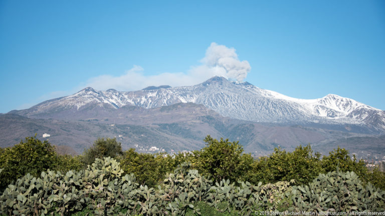 Broad snow-streaked Mount Etna with a small summit plume, seen beyond prickly pear and green foreground vegetation.