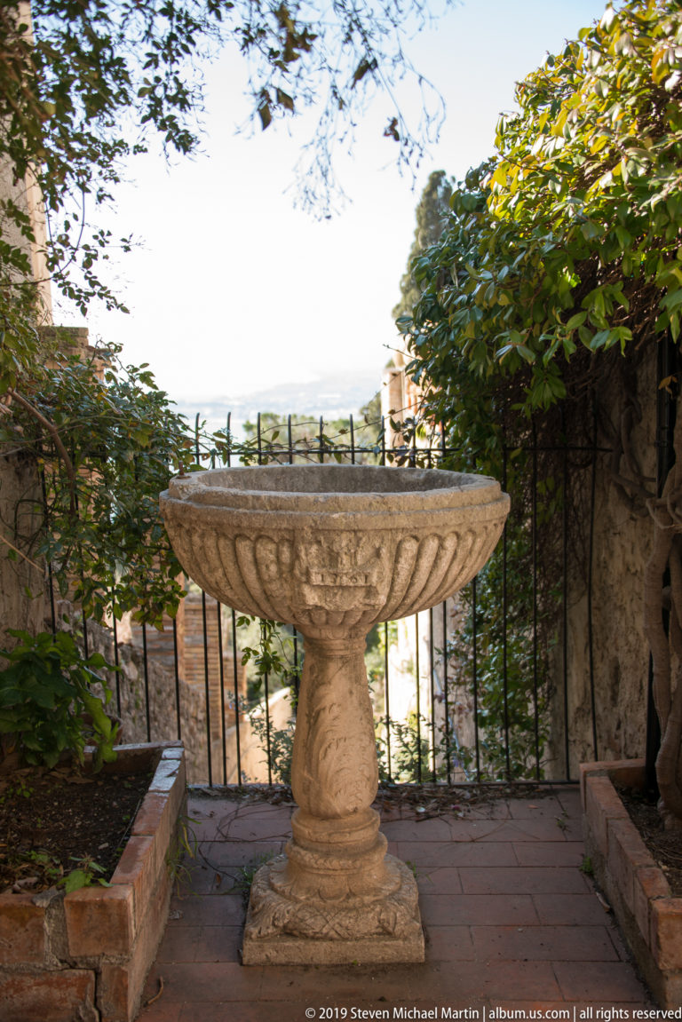 Weathered fluted stone basin on a carved pedestal beside an iron railing, trees, and a distant coastal view.