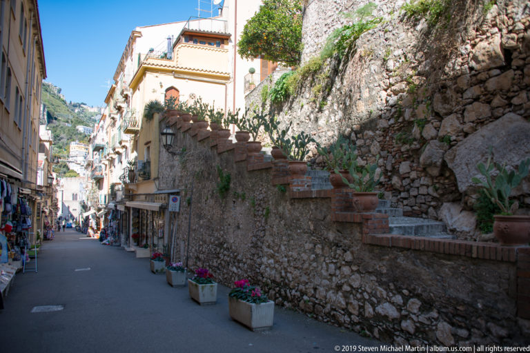 Narrow street beside a tall rough-stone wall with brick edging, cactus, planters, shops, and hillside buildings beyond.