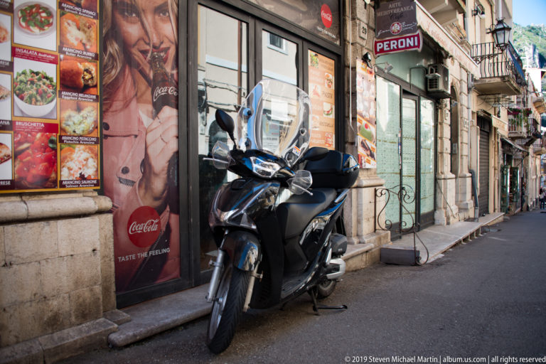 Black motor scooter parked beside restaurant menu boards and beverage advertisements on a narrow Taormina street.