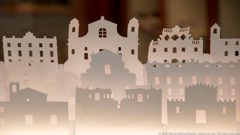 Layered white and gray cutout silhouettes of churches, towers, buildings, and ruins arranged as a decorative skyline.