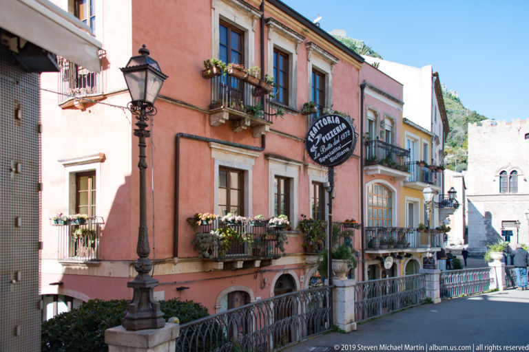 Pink historic building with flowered balconies, wrought-iron railings, streetlamp, and hanging trattoria sign on a Taormina street.