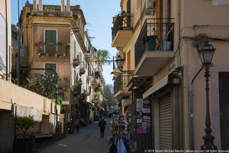 Narrow street lined with balconies, shutters, lamps, souvenir displays, and pedestrians, with palms visible farther ahead.