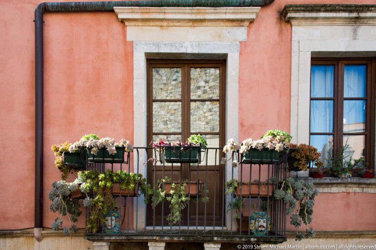 Flower-filled wrought-iron balcony between dark wooden windows on a salmon-colored stucco façade with pale stone trim.